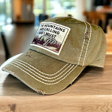 Load image into Gallery viewer, An olive green baseball cap with white stitching on crown and bill. It has a white fabric patch with a mountain scene that says, The mountains are calling and I must go.&quot; The edged of the patch are frayed. 