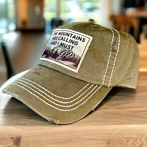 An olive green baseball cap with white stitching on crown and bill. It has a white fabric patch with a mountain scene that says, The mountains are calling and I must go.