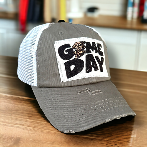 Gray and white mesh low profile and unstructured trucker hat with a distressed fabric patch that says Game Day with a leopard print lightening strike that takes the place of the letter A in Game.