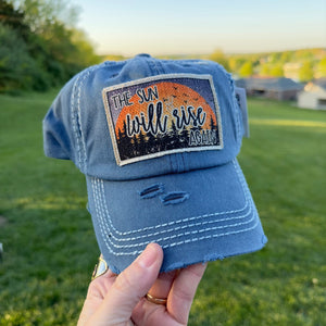 Blue full cotton baseball hat with a frayed edge fabric patch that says, the sun will rise again.