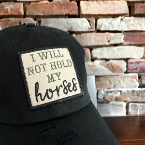 black mesh baseball hat with an embroidered patch that says, &quot;I will not hold my horses&quot;. The patch border is black and the words are olive green and black.