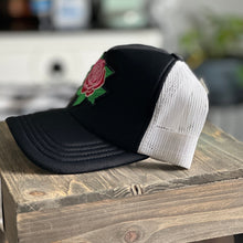 Load image into Gallery viewer, Black front and white mesh back mid profile trucker hat with an embroidered red and pink rose with green leaves patch. The patch has distressed edges.