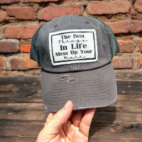 Dark Gray Trucker hat, with a rectangle shaggy edge white patch that says, The best things in life mess up your hair. the patch has a thin black border and frayed edges.