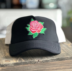 Black front and white mesh back mid profile trucker hat with an embroidered red and pink rose with green leaves patch. The patch has distressed edges.