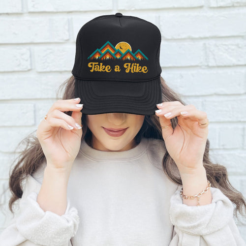 A woman wearing a Black high profile foam trucker hat with a colorful mountain image with the words, Take a Hike in yellow retro font