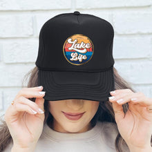 Load image into Gallery viewer, A woman wearing this black high profile rope trucker hat with a colorful round Lake LIfe graphic printed on the front. Lake life is printed in white cursive bubble font. The colors in the background are in a strip pattern. Blues, yellows, reds