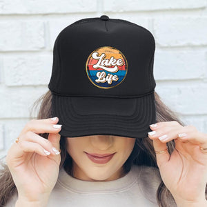 A woman wearing this black high profile rope trucker hat with a colorful round Lake LIfe graphic printed on the front. Lake life is printed in white cursive bubble font. The colors in the background are in a strip pattern. Blues, yellows, reds
