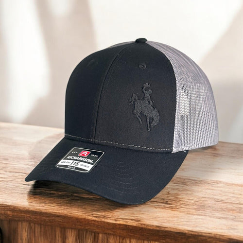 Black and charcoal mesh Richardson Low Pro 112 Trucker hat with an embroidered cowboy riding a bucking bronco. The thread is black making it tone on tone.