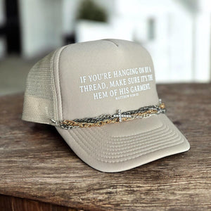 Tan trucker hat with the phrase, If you&#39;re hanging on by a thread, make sure it&#39;s the hem of His garment. matthew 9:20-22. It in white font