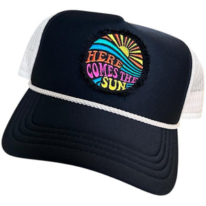 A black mid profile trucker hat with a cream rope and cream mesh. It has an embroidered here comes the sun round raggy patch. The image is embroidered onto a black frayed fabric using bright colors of orange pink, yellow, blue and teal.