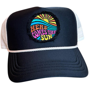 A black mid profile trucker hat with a cream rope and cream mesh. It has an embroidered here comes the sun round raggy patch. The image is embroidered onto a black frayed fabric using bright colors of orange pink, yellow, blue and teal.