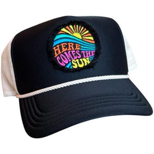 A black mid profile trucker hat with a cream rope and cream mesh. It has an embroidered here comes the sun round raggy patch. The image is embroidered onto a black frayed fabric using bright colors of orange pink, yellow, blue and teal.
