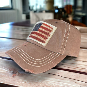 Vintage washed brown cap with some distressing and white stitching. I has a rustic American flag printed on tan frayed fabric and is securely attached to the hat. The hat has a curved bill.